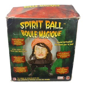May include: A Spirit Ball Boule Magique product box featuring a graphic of a head wearing an orange turban inside a glass sphere on a black stand. The box includes text such as Eerie lighting, Eyes light up, Sound Activated, and 14 inch mysterious globe.