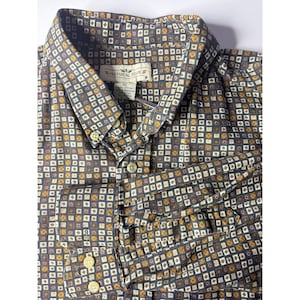 Puede incluir: Camisa abotonada con un patrón de cuadrados repetidos en tonos azules, marrones y dorados. La camisa tiene cuello, puños abotonados y la etiqueta dice "NATURAL ISSUE". La camisa es talla XL.