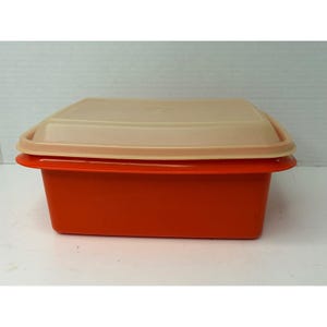 Può includere: Un contenitore rettangolare con base arancione brillante e coperchio beige chiaro. Il contenitore presenta una superficie liscia e piatta con un bordo leggermente rialzato nella parte superiore, che garantisce una chiusura sicura del coperchio.