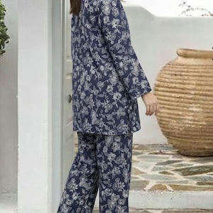 Könnte beinhalten: Ein zweiteiliges Loungewear-Set bestehend aus einer langärmeligen Tunika und einer passenden Hose mit geradem Bein. Das Kleidungsstück ist marineblau mit einem durchgehenden, weißen, floralen Linienmuster. Das Oberteil hat einen runden Ausschnitt.