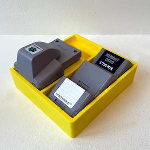Może przedstawiać: Jaskrawożółta plastikowa taca do przechowywania zawierająca trzy szare akcesoria do konsoli Nintendo 64. Przedmioty obejmują szary expansion pak z logo Nintendo 64, szarą kartę pamięci z napisem 256KB oraz szary controller pak z białą etykietą NINTENDO 64.