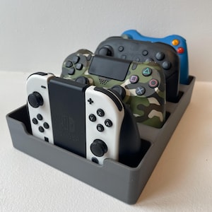 Könnte beinhalten: Eine graue, rechteckige Aufbewahrungsschale mit drei Videospiel-Controllern. Der vordere Controller ist weiß und schwarz mit dem Switch-Logo. Dahinter befindet sich ein Controller mit Tarnmuster, gefolgt von einem schwarzen und einem blauen Controller.