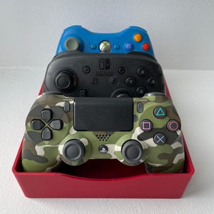 May include: A red tray holds three video game controllers: a blue Xbox controller, a black Nintendo Switch controller, and a camouflage-patterned PlayStation controller. The controllers are stacked on top of each other.