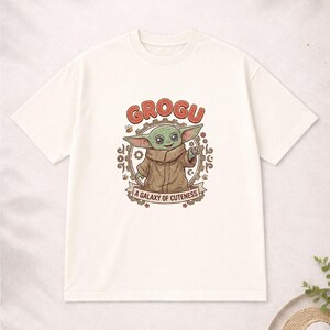 May include: Off-white t-shirt with a Grogu cartoon design. The design features the word "GROGU" in red, a circular frame, and the text "A GALAXY OF CUTENESS" on a banner. A casual wear item.