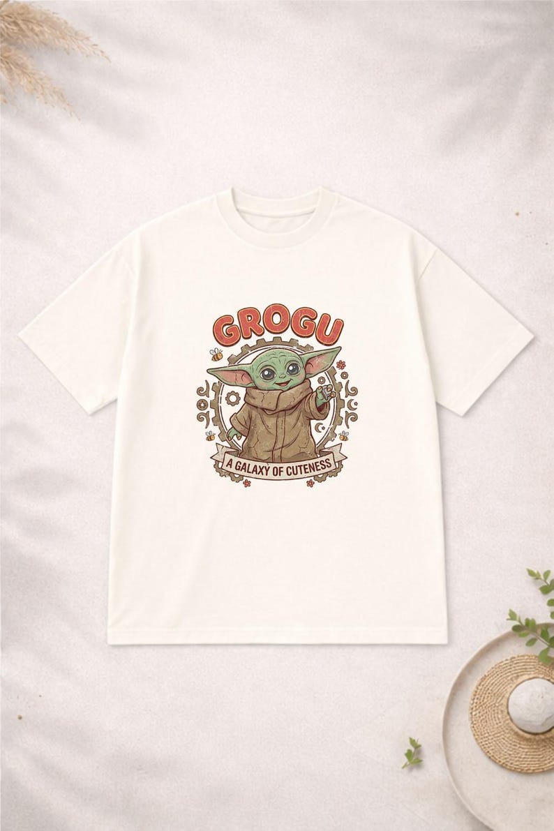 May include: Off-white t-shirt with a Grogu cartoon design. The design features the word "GROGU" in red, a circular frame, and the text "A GALAXY OF CUTENESS" on a banner. A casual wear item.