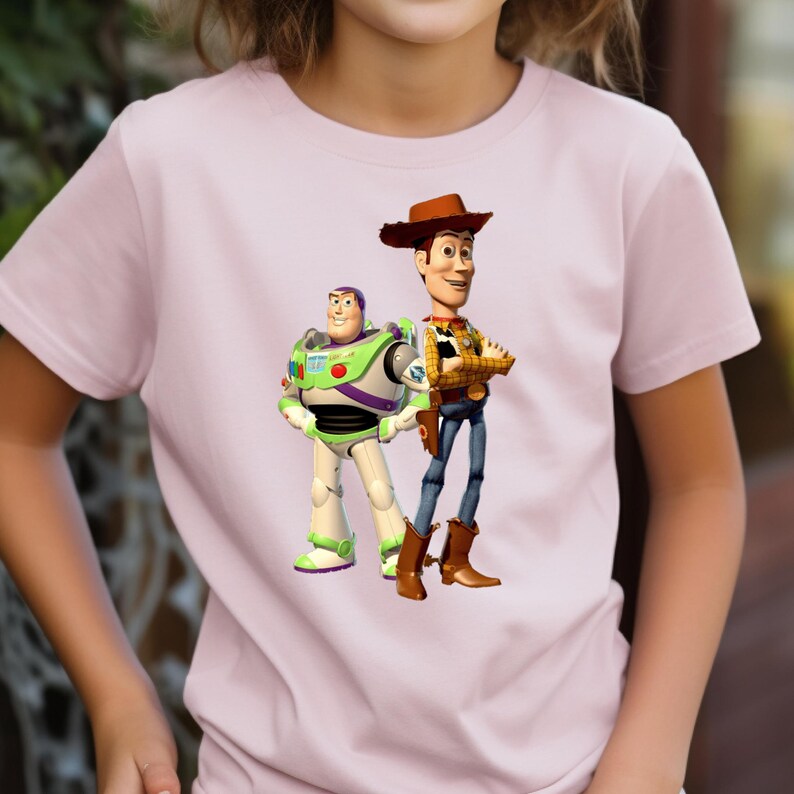 May include: A light pink short-sleeved t-shirt featuring a graphic print of the characters Buzz Lightyear and Woody from Toy Story. Buzz Lightyear is depicted in his white, green, and purple space suit, while Woody wears a cowboy hat, yellow shirt.