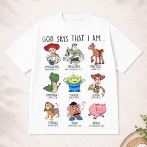 May include: White t-shirt featuring cartoon characters with the text "GOD SAYS THAT I AM...". Each character has a corresponding word like "Empowered" and a Bible verse reference. The shirt has a crew neck and short sleeves.