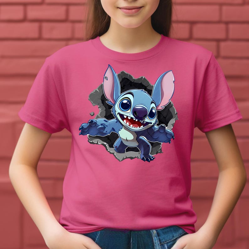 May include: A pink t-shirt with a cartoon image of Stitch, a blue alien, breaking through a hole. Stitch has large ears and a wide smile. The background is a brick wall.