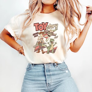 May include: Cream-colored t-shirt featuring a vintage-style graphic of Toy Story characters. The design includes Woody, Buzz Lightyear, Rex, and the Toy Story logo with the text "Since 1995".
