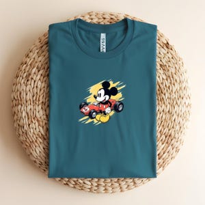 May include: Teal t-shirt with a cartoon of Mickey Mouse in a red race car against a yellow backdrop. The shirt is presented on a circular woven mat.