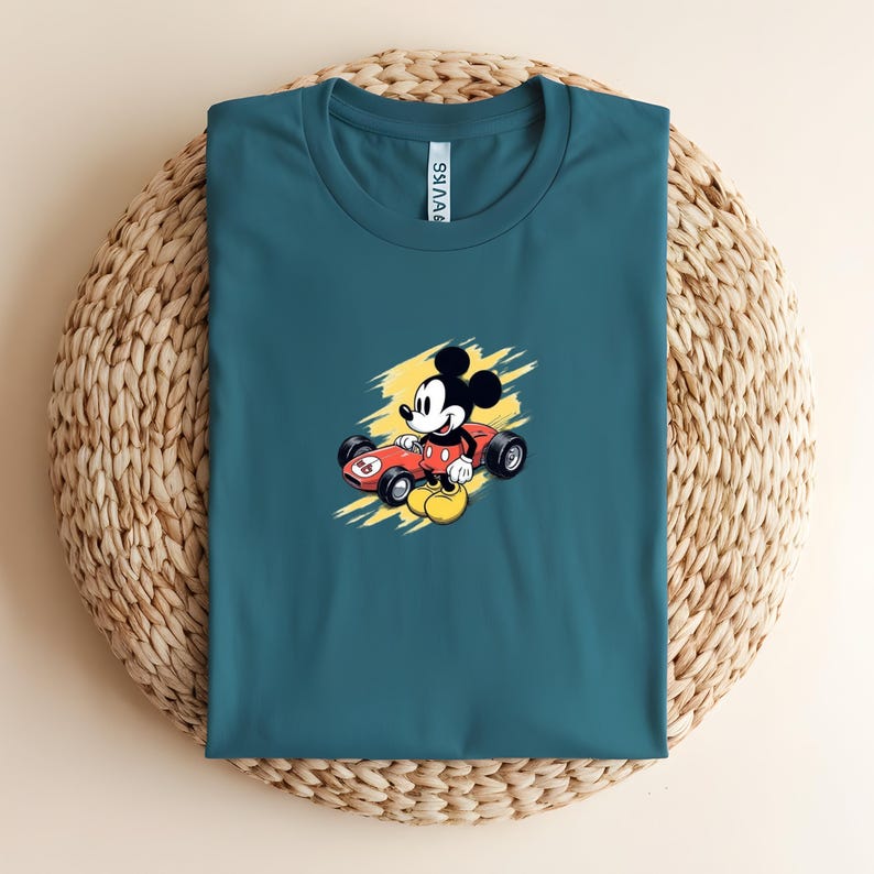 May include: Teal t-shirt with a cartoon of Mickey Mouse in a red race car against a yellow backdrop. The shirt is presented on a circular woven mat.