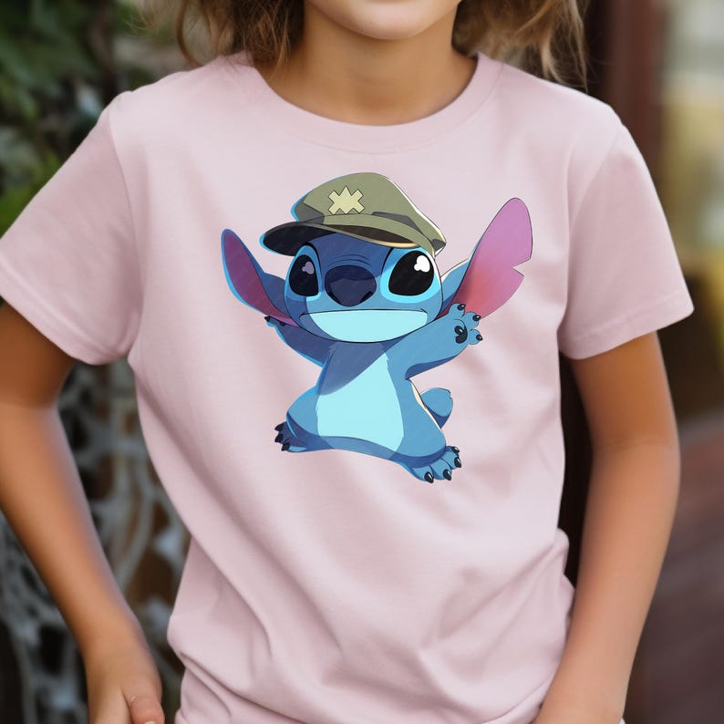 May include: A light pink short-sleeved t-shirt featuring a graphic print of a blue alien character wearing a tan military-style cap with a yellow star emblem. The character has large ears, a round nose, and is depicted with arms outstretched.