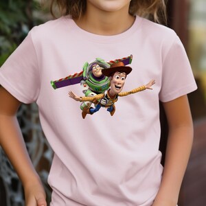 May include: A light pink t-shirt featuring a graphic of Woody and Buzz Lightyear from the animated movie Toy Story. Woody is wearing a cowboy hat and Buzz has his wings extended. The characters appear to be flying.