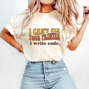 May include: Cream-colored t-shirt with the text "I CAN'T FIX YOUR PRINTER I write code." in retro orange and yellow lettering. The shirt is paired with light blue denim jeans. A casual, everyday wear top.