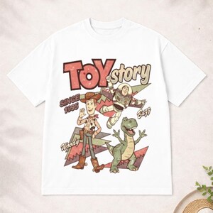 May include: White t-shirt featuring a retro-style graphic of Toy Story characters Woody, Buzz Lightyear, and Rex. The words "Toy Story" and "Since 1995" are also printed on the shirt. A fun, nostalgic design for fans of the animated film.