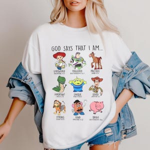 May include: White t-shirt featuring cartoon characters with the text "GOD SAYS THAT I AM..." and words like "Empowered," "Treasured," and "Precious." The shirt has a crew neck and short sleeves. The design includes various characters and associated Bible verses.