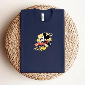 May include: Navy blue t-shirt with a cartoon Mickey Mouse graphic in a red race car against a yellow backdrop. The shirt is folded on a circular woven mat. The shirt has a crew neck and a small white tag.