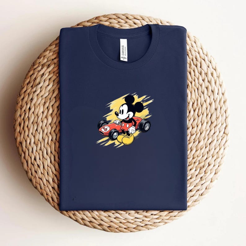 May include: Navy blue t-shirt with a cartoon Mickey Mouse graphic in a red race car against a yellow backdrop. The shirt is folded on a circular woven mat. The shirt has a crew neck and a small white tag.