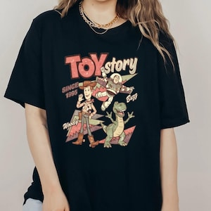May include: Black t-shirt featuring a graphic of characters from the movie Toy Story. The design includes Woody, Buzz Lightyear, and Rex, with the text "Toy Story Since 1995". The design is in shades of red, pink, green, and white.