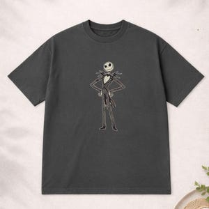Puede incluir: Camiseta gris oscuro con una ilustraci&oacute;n de dibujos animados de Jack Skellington de Pesadilla antes de Navidad. El personaje est&aacute; representado con su traje caracter&iacute;stico en blanco y negro con los brazos en jarras. La camiseta es de un material suave.