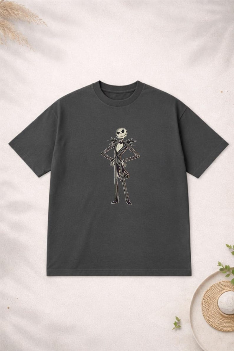 Puede incluir: Camiseta gris oscuro con una ilustraci&oacute;n de dibujos animados de Jack Skellington de Pesadilla antes de Navidad. El personaje est&aacute; representado con su traje caracter&iacute;stico en blanco y negro con los brazos en jarras. La camiseta es de un material suave.