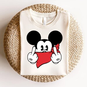 May include: Cream-colored t-shirt featuring a cartoon mouse with black ears and a red bandana. The mouse is giving the middle finger with both hands. The shirt is folded on a woven surface.