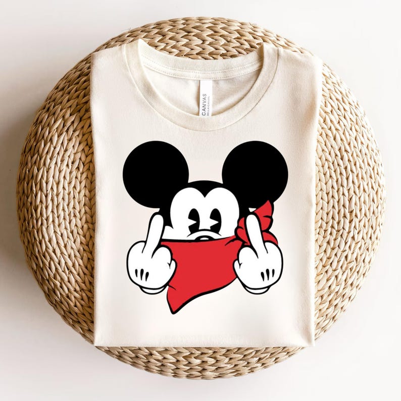 May include: Cream-colored t-shirt featuring a cartoon mouse with black ears and a red bandana. The mouse is giving the middle finger with both hands. The shirt is folded on a woven surface.