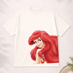 Puede incluir: Camiseta de color crema con una ilustraci&oacute;n colorida de Ariel de La Sirenita. Ariel tiene el pelo largo rojo, ojos azules y una expresi&oacute;n sonriente. La camiseta es de estilo cuello redondo cl&aacute;sico.