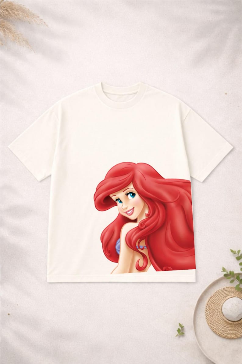 Puede incluir: Camiseta de color crema con una ilustraci&oacute;n colorida de Ariel de La Sirenita. Ariel tiene el pelo largo rojo, ojos azules y una expresi&oacute;n sonriente. La camiseta es de estilo cuello redondo cl&aacute;sico.