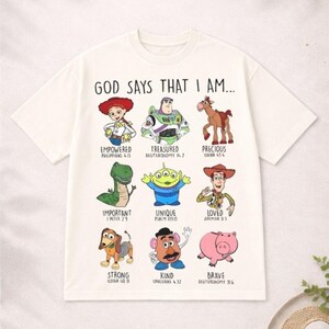 May include: Off-white t-shirt featuring cartoon characters with the text "GOD SAYS THAT I AM..." and various positive affirmations. The shirt includes characters like Woody, Buzz Lightyear, and others, with corresponding Bible verses.