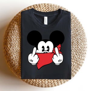 May include: Black t-shirt featuring a cartoon mouse with black ears, white gloves, and a red bandana. The mouse is giving the middle finger with both hands. The shirt is folded on a woven surface.