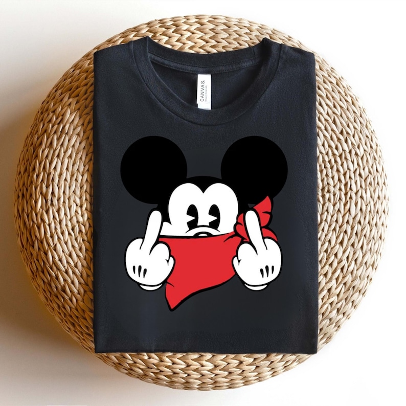 May include: Black t-shirt featuring a cartoon mouse with black ears, white gloves, and a red bandana. The mouse is giving the middle finger with both hands. The shirt is folded on a woven surface.