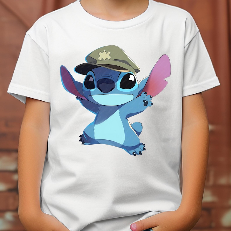 May include: A white short-sleeved t-shirt featuring a graphic print of a blue alien character wearing a tan military-style cap with a yellow cross emblem. The character has large ears, a wide smile, and is depicted in a dynamic pose.