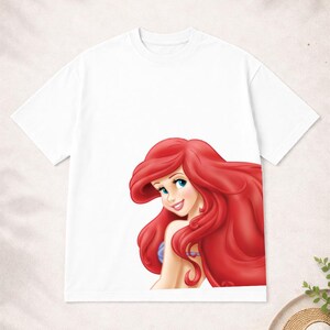 Puede incluir: Camiseta blanca con una ilustraci&oacute;n de dibujos animados de un personaje con pelo largo rojo y ojos azules. El personaje est&aacute; sonriendo. La camiseta est&aacute; hecha de un material suave y c&oacute;modo.