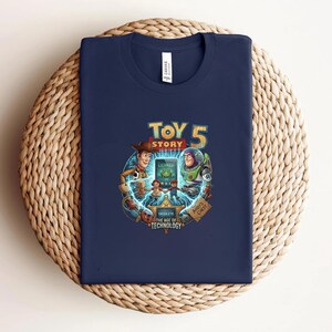 May include: Navy blue t-shirt featuring a colorful graphic from the movie Toy Story 5. The design includes Woody, Buzz Lightyear, and other characters. The text "TOY STORY 5" is prominently displayed, along with the text "OBSOLETE THE AGE OF TECHNOLOGY".