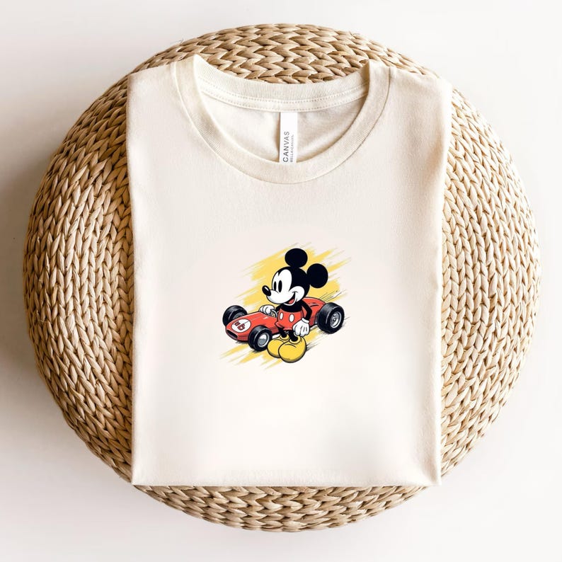 May include: Cream-coloured t-shirt featuring a cartoon image of Mickey Mouse driving a red race car. The graphic has a yellow background. The shirt is folded on a woven, circular surface.