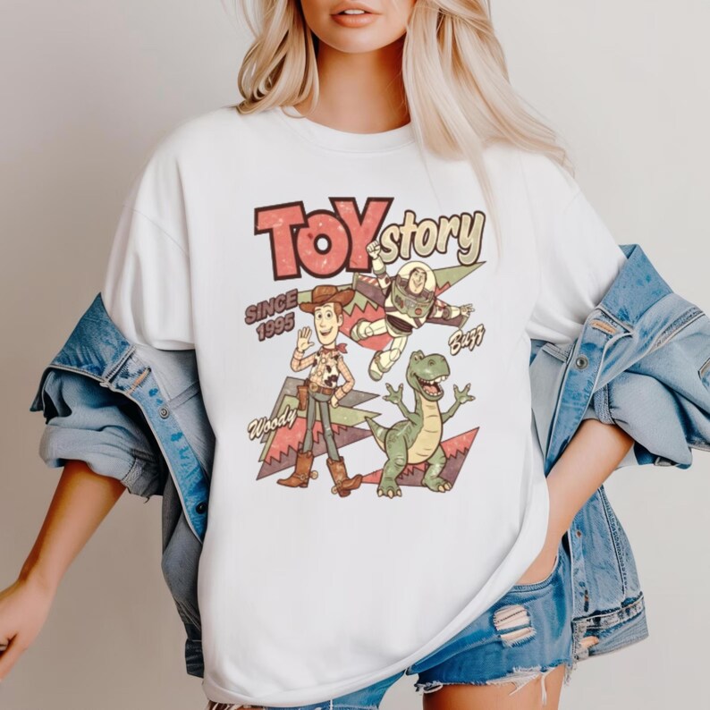 May include: White t-shirt featuring a retro-style graphic of Toy Story characters, including Woody, Buzz Lightyear, and Rex. The design includes the text "Toy Story Since 1995". The shirt has a classic crew neck and short sleeves.