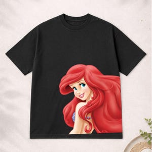 Puede incluir: Camiseta negra de manga corta con una colorida ilustraci&oacute;n de Ariel de La Sirenita. Ariel tiene el pelo largo y rojo y est&aacute; sonriendo. La camiseta est&aacute; hecha de un material suave.