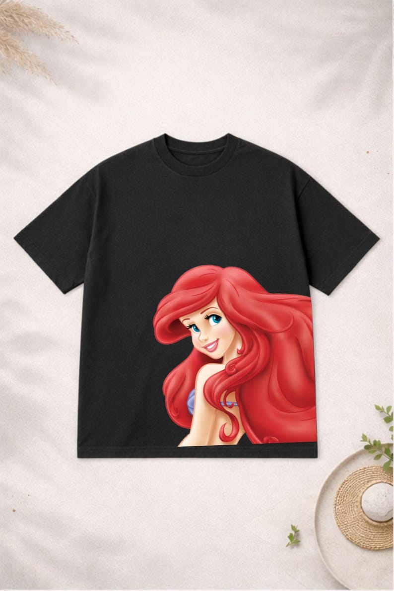 Puede incluir: Camiseta negra de manga corta con una colorida ilustraci&oacute;n de Ariel de La Sirenita. Ariel tiene el pelo largo y rojo y est&aacute; sonriendo. La camiseta est&aacute; hecha de un material suave.