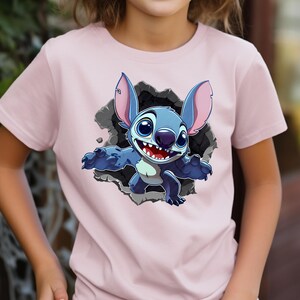 May include: A pale pink t-shirt with a cartoon image of Stitch bursting through a grey wall. Stitch is blue with large ears, big eyes, and a wide smile. The t-shirt is made of a soft material and is suitable for children.