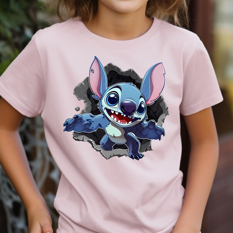 May include: A pale pink t-shirt with a cartoon image of Stitch bursting through a grey wall. Stitch is blue with large ears, big eyes, and a wide smile. The t-shirt is made of a soft material and is suitable for children.