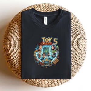 May include: Black t-shirt featuring a colorful graphic from Toy Story 5. The design includes Woody, Buzz Lightyear, and other characters, with the text "TOY STORY 5" and "OBSOLETE THE AGE OF TECHNOLOGY". The shirt is folded on a woven surface.
