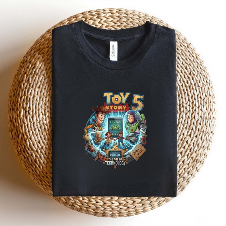 May include: Black t-shirt featuring a colorful graphic from Toy Story 5. The design includes Woody, Buzz Lightyear, and other characters, with the text "TOY STORY 5" and "OBSOLETE THE AGE OF TECHNOLOGY". The shirt is folded on a woven surface.