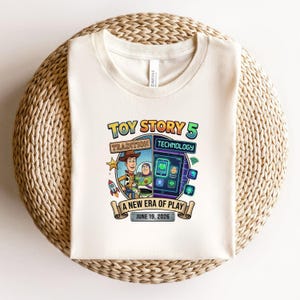 May include: Cream-colored t-shirt featuring a colorful graphic for "Toy Story 5." The design includes Woody and Buzz Lightyear, a tablet with app icons, and the text "A New Era of Play, June 19, 2026."