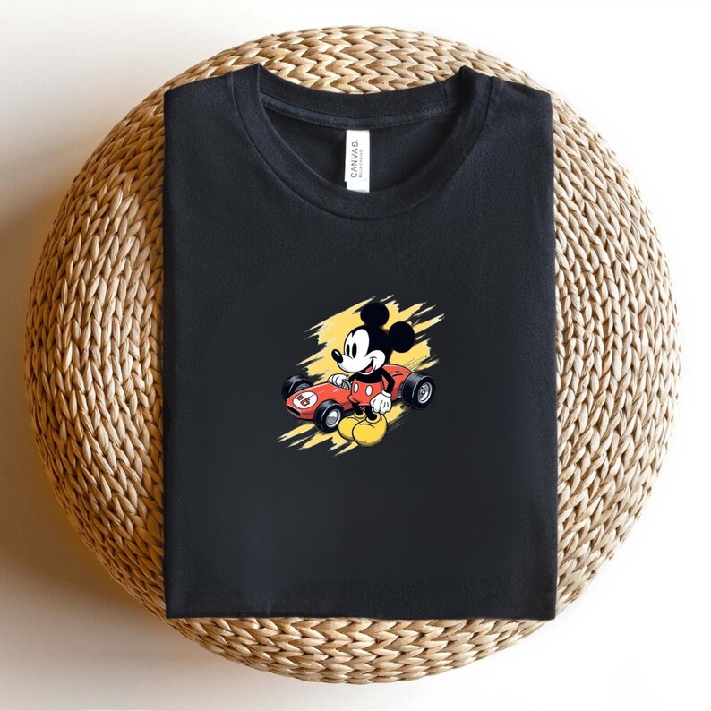 May include: Black t-shirt with a cartoon Mickey Mouse driving a red racing car, number 8 on the side. The graphic is set against a yellow background with black and white accents. The shirt is folded on a woven surface.