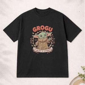 May include: Black t-shirt with a Grogu graphic. The design features the word "GROGU" in orange, a cartoon image of Grogu, and the phrase "A Galaxy of Cuteness". The t-shirt has a crew neck.