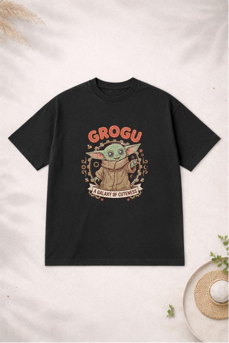 May include: Black t-shirt with a Grogu graphic. The design features the word "GROGU" in orange, a cartoon image of Grogu, and the phrase "A Galaxy of Cuteness". The t-shirt has a crew neck.