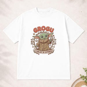 May include: White t-shirt with a Grogu graphic. The design features the word "GROGU" in red, an illustration of Grogu, and the phrase "A GALAXY OF CUTENESS" on a banner. The shirt has a crew neck.