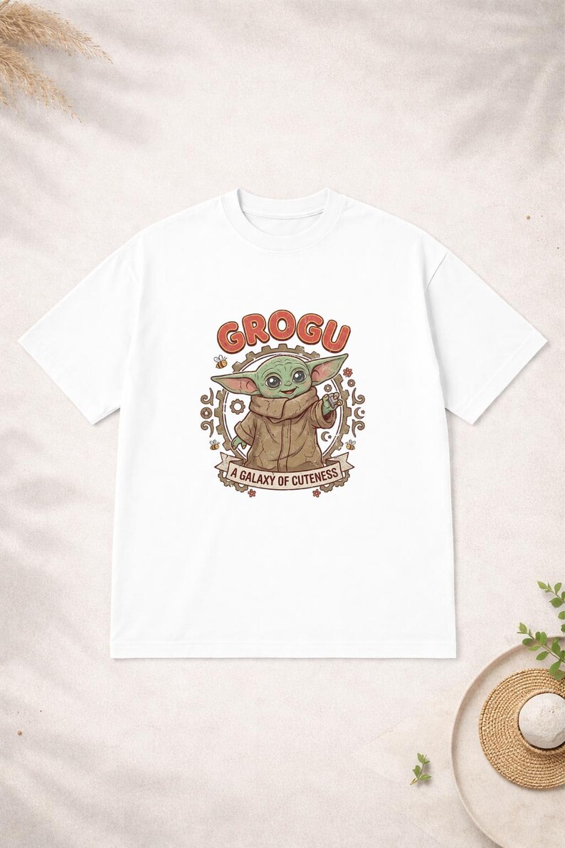 May include: White t-shirt with a Grogu graphic. The design features the word "GROGU" in red, an illustration of Grogu, and the phrase "A GALAXY OF CUTENESS" on a banner. The shirt has a crew neck.
