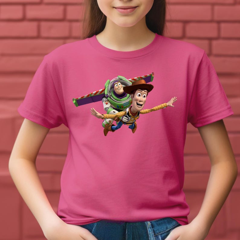 May include: A pink t-shirt featuring a graphic of Woody and Buzz Lightyear from Toy Story. Woody is wearing a cowboy hat and Buzz has his wings extended. The characters are depicted mid-flight, with arms outstretched.